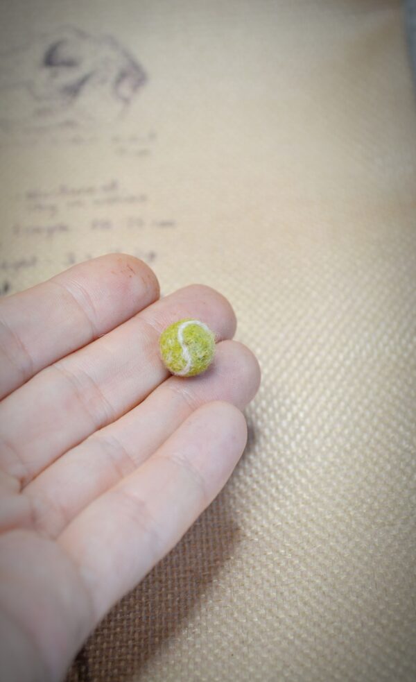 Miniature wool tennis ball, handmade with curved white lines. A tiny felted accessory for dollhouses, pets, or sports-themed mini scenes.